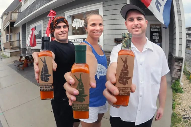 Hank Sauce's Matt Pittaluga (left) with Kaitlin and Brian "Hank" Ruxton, and three of the company's hot-sauce varieties. Brian Ruxton comes up with the recipes; Pittaluga designed the logo and square bottle. A third cofounder, Josh Jaspan, handles the business end. (DAVE GRIFFIN / For The Inquirer)
