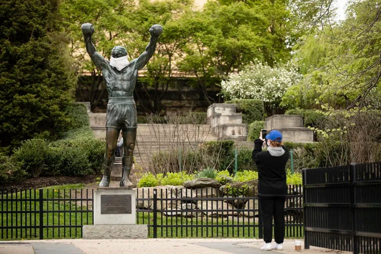 A person wearing a protective face mask as a precaution against the coronavirus makes a photograph of the Rocky statue outfitted with mock surgical face mask at the Philadelphia Art Museum on Tuesday.