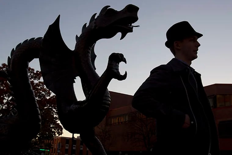 Drexel senior Arsen Nikiforouk is silhouetted as he walks by the statue of Mario the Magnificent, the Drexel Dragon, at 33rd and Market Streets as the sun sets. ( CHARLES FOX / Staff Photographer )