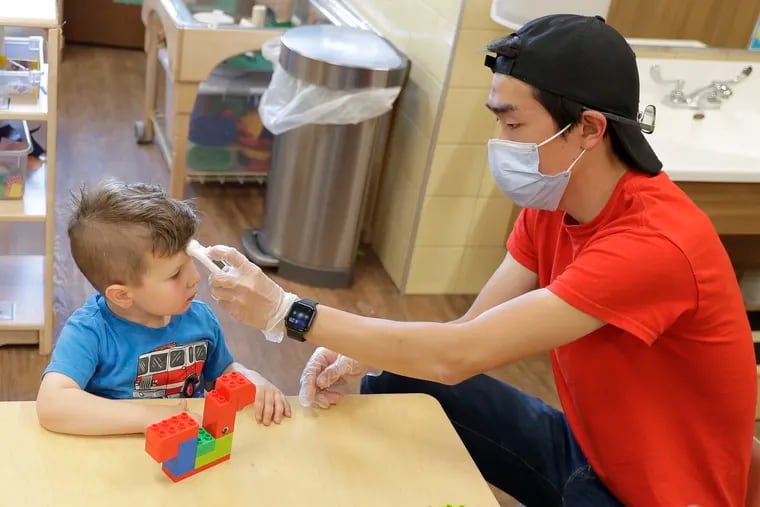 In this May 27, 2020, photo, Aaron Rainboth, a teacher at the Frederickson KinderCare day-care center in Tacoma, Wash., wears a mask as he takes the temperature of Benjamin Simpson, 4, after he complained of feeling hot following an outdoor play period but found it to be normal.