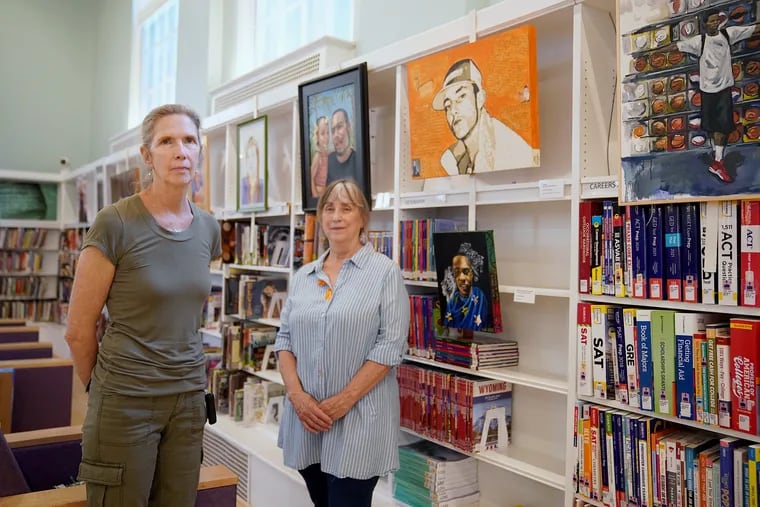 Laura Madeleine (left), executive director of the Souls Shot Portrait Project, and artist Ann Price Hartzell next to Hartzell's painting of Adam Hammer (in orange), who was shot and killed in 2004, at the Logan Library in North Philadelphia on Wednesday, May 26, 2021. The library just finished exhibiting portraits from the Souls Shot project, which connects artists with families of gun violence victims to memorialize those killed.