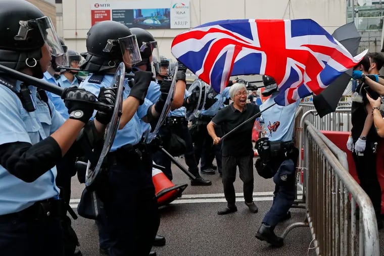 In this Wednesday, June 12, 2019, photo, a woman waves a British flag as policemen in anti-riot gear stand guard against the protesters on a closed-off road near the Legislative Council in Hong Kong. China promised that for 50 years after Britain gave up control of its last colony in 1997, this shimmering financial enclave would get to keep freedoms absent in the communist-ruled mainland that many here don't want to live without. (AP Photo/Kin Cheung)