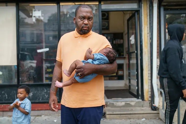Oronde McClain holds his 2-month-old son, Jalen, while his 3-year-old son, Jayce, explores. Lano Lewis (far right), a student McClain is mentoring, stands by on the East Mount Airy block where McClain was shot as a child.