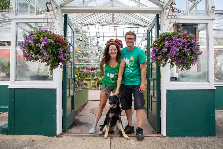 Stacey and Sean McNicholl of GreenHorn Gardens in Upper Darby, along with Rebel, their 5-month-old German shepherd, stand in front of the entrance of their greenhouse at Arlington Cemetery.