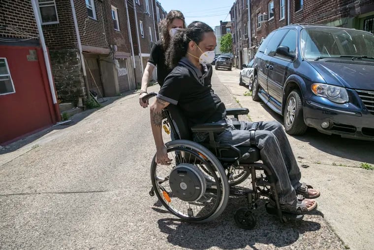 Shaylin Sluzalis helps Germán Parodi after they pose for a portrait outside of their Juniata Park home. Philadelphia has been slow to provide homebound vaccinations for people. Parodi, who is  quadriplegic, had to rely on the Black Doctors COVID Consortium to get a shot.