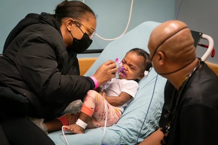 "Tyshanek Sutton (left) places a nebulizer mask over the mouth and nose of her daughter, Zariya Sutton-Pack, with help from John Smith (right), the only respiratory therapist on duty in the emergency department at St. Christopher’s Hospital for Children."