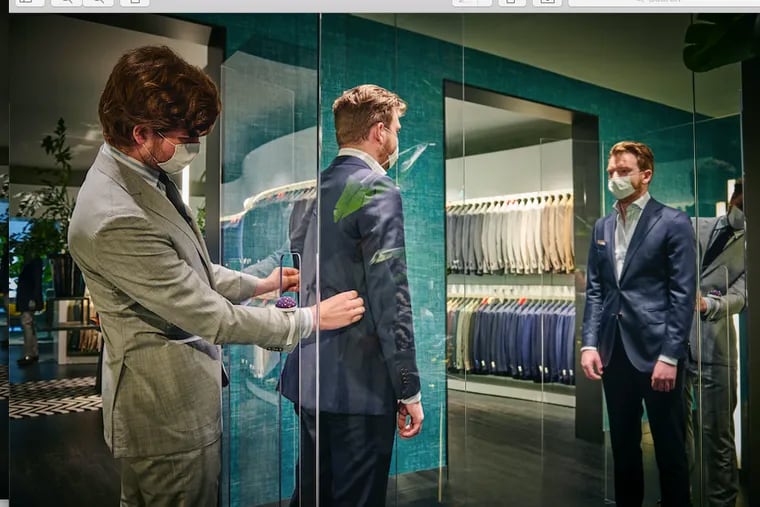 To get fitted for a suit at Suitsupply during the coronavirus pandemic, a customer is surrounded by three 8- to 9-foot clear plastic panels. A tailor reaches through a cutout in a panel to hem and pin the suit.