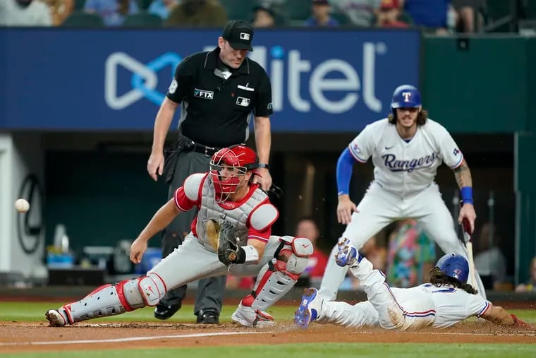 Philadelphia Phillies catcher J.T. Realmuto waits on the throw to the plate as Texas Rangers' Josh Smith scores on a single by Brad Miller in the second inning of a baseball game, Wednesday, June 22, 2022, in Arlington, Texas. Jonah Heim, right rear, who also scored on the single, and home plate umpire Chris Conroy, left rear, look on at the play,. (AP Photo/Tony Gutierrez)
