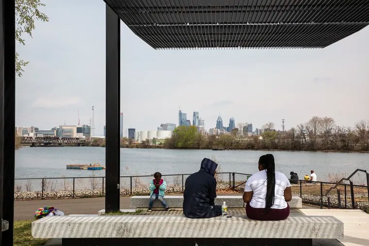 People sit out before the Schuylkill River with a view of the Philadelphia skyline at Bartram's Garden in Philadelphia on a calm day in April.