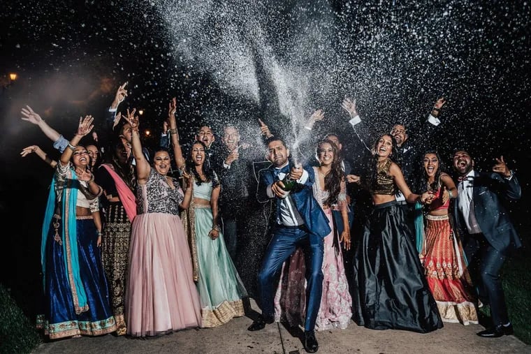 In front of a colorfully clad bridal party and Rita Shah, groom Krshna Patel pops a just-shaken bottle of champagne and a million tiny white bubbles are frozen against a backdrop of dark sky.