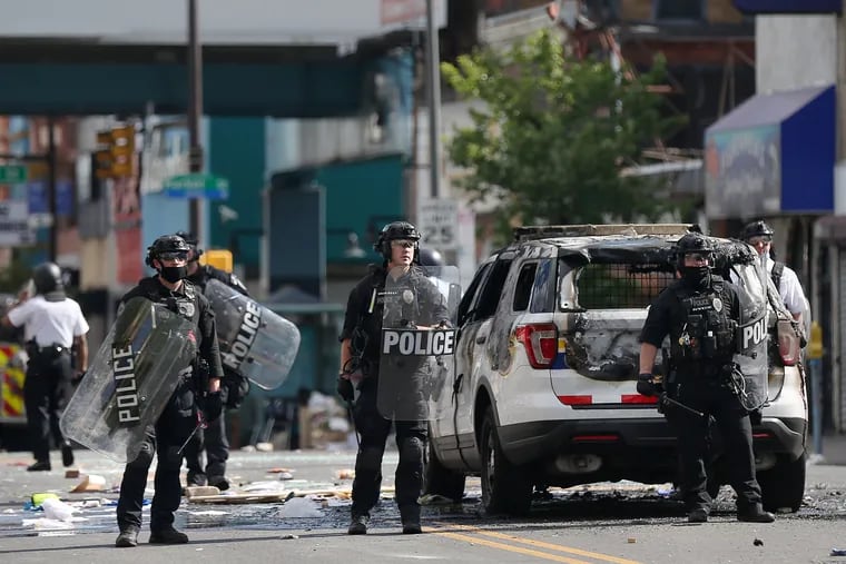 University of Pennsylvania Police Department officers in tactical gear stand in front of a burned-out Philadelphia Police cruiser as they face off against a crowd on 52nd Street between Arch and Market Streets in West Philadelphia on Sunday, May 31, 2020.