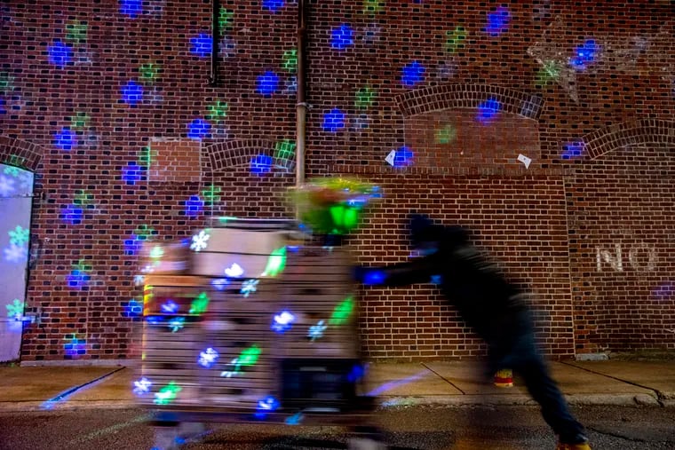December 21, 2020: A worker from the 9th Street Market in South Philadelphia pushes boxes of produce to a refrigerated storage area, passing through projected light snowflakes on a wall.