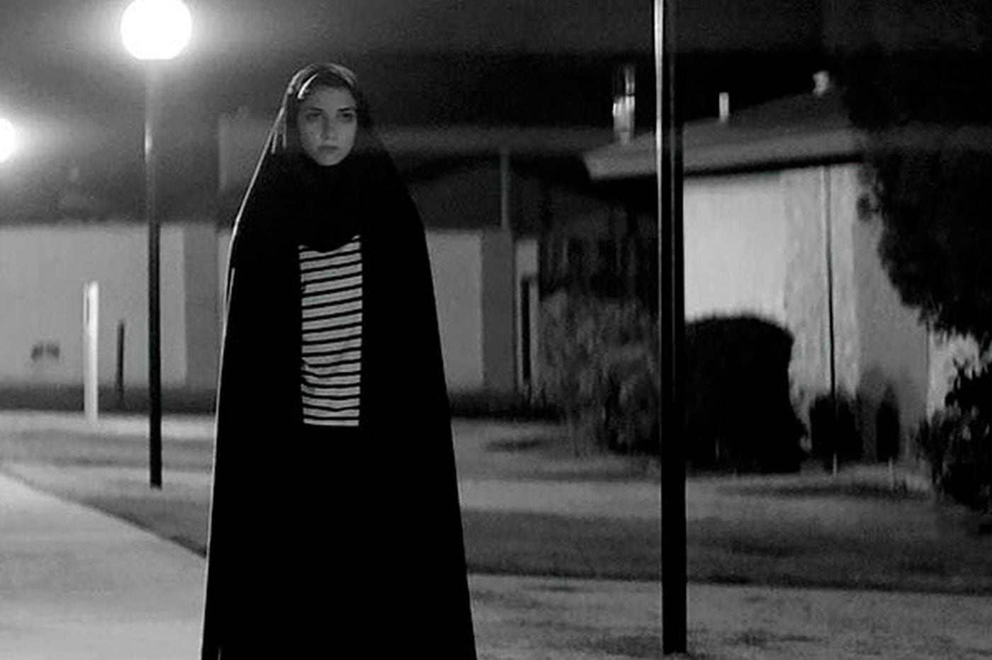 Image is from the film 'A Girl Walks Home Alone at Night' (2014). In an empty street, a young woman wearing a long coat and headscarf walks alone at night.