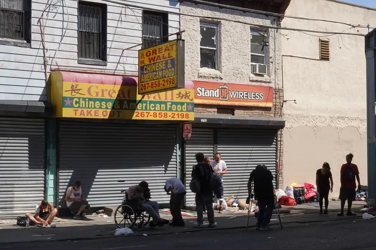 A group of people gather along the sidewalk in front of shutter storefronts, 2700 block of Kensington Ave., Philadelphia on Thursday, June 27, 2019. This section of the city is struggling with drug abuse and poverty.