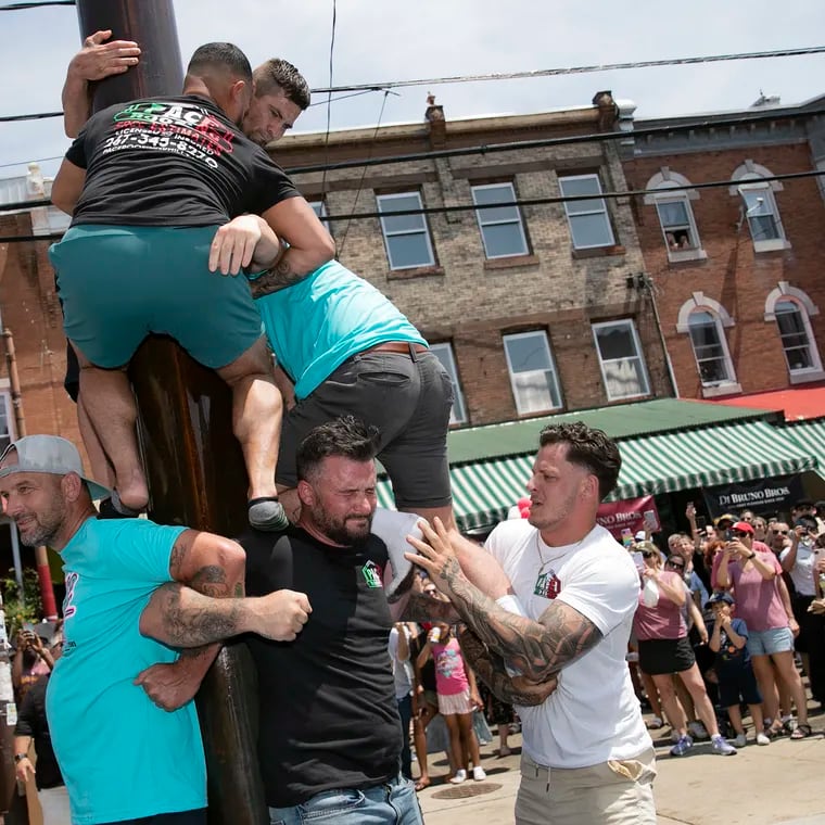 A group of men with Pace Roofing LLC compete in the grease pole contest at the South 9th Street Italian Market Festival in Philadelphia on Saturday, May 21, 2022. They won on their second attempt to climb the pole and grab the meat and cheese hanging on top.