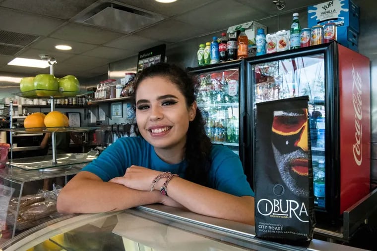 Isabella Bacciarini, a junior at String Theory High School and an employee at Logan Square Cafe in Center City, next to a bag of Obura brand coffee beans at the cafe.
