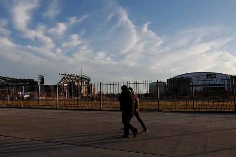 Pedestrians walk along the sidewalk on Pattison Avenue in South Philadelphia with the Wells Fargo Center and Lincoln Financial Field in the background on Sunday, December 19, 2021.