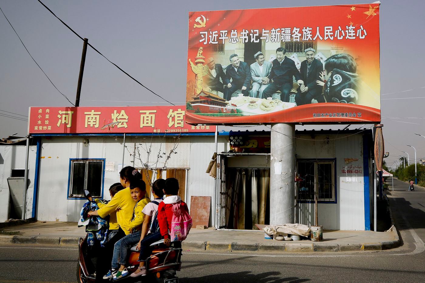 In this Sept. 20, 2018, photo, an Uighur woman uses an electric-powered scooter to fetch schoolchildren as they ride past a picture showing China's President Xi Jinping joining hands with a group of Uighur elders at the Unity New Village in Hotan, in western China's Xinjiang region. 