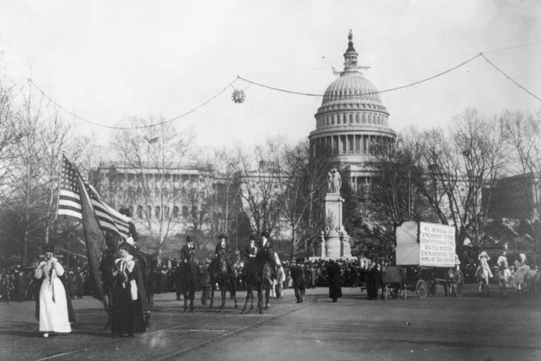 Women on horseback marched in a parade behind flag-bearers on Pennsylvania Ave. A horse and cart pulled a large sign, "We demand an amendment to the constitution of the United States enfranchising the women of this country."