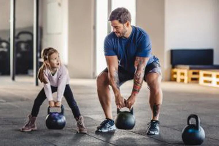 Father doing his training with kettlebells in gym while his little daughter supporting him.