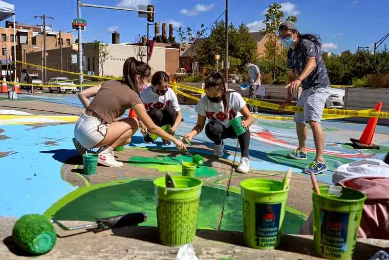 Artist Chenlin Cai (standing) gives instructions to volunteers Mariana Di (left), and Kappa Phi Lambda sorority sisters from Drexel Univ. Nina Wang (center) and Athena Le (right) as they repaint a mural at 10th and Vine Streets Sept. 26, 2021. Cai painted the original mural two years ago and designed the new version, as the neighborhood looks to create a play space for children in Chinatown.