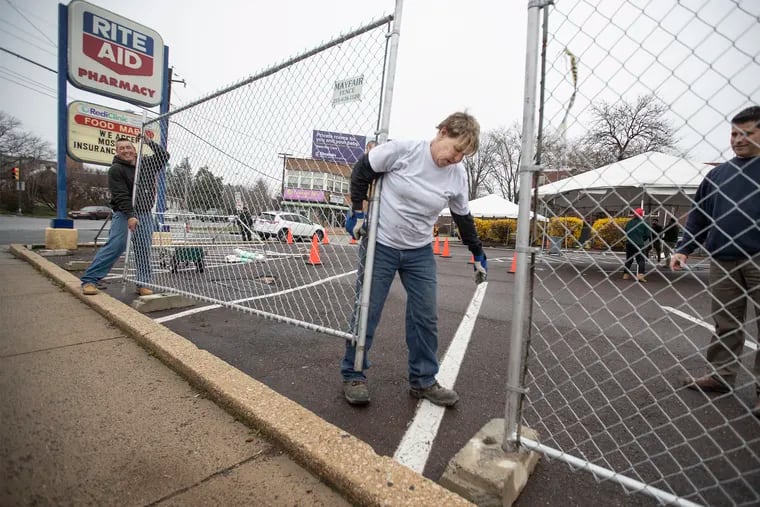 Joan Twilley (center) and Clint Twilley (left) erect a chain-link fence around a Rite Aid parking lot on the corner of Stenton Avenue and Washington Lane, where a private coronavirus temporary testing site is being built, on Thursday.