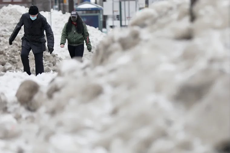 Pedestrians walk past high piles of snow created by plow trucks along Market Street at Schuylkill Avenue in University City. That pile might get a whitewashing Sunday.