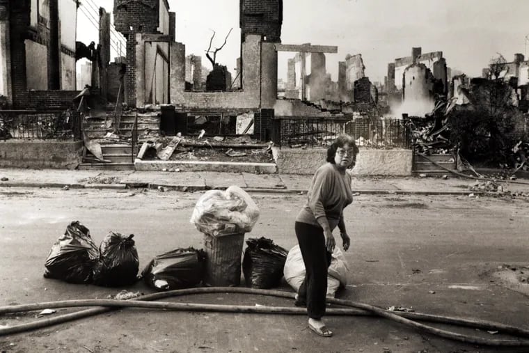 On May 15, 1985, trash day in the neighborhood, resident Myrna Zachary of the 6200 block of Pine St. puts out her garbage across from the destroyed homes. Her home received only minor damage in the bombing and resulting fire that killed eleven MOVE victims. Fire hoses still run down street as smoke still rises from rubble.