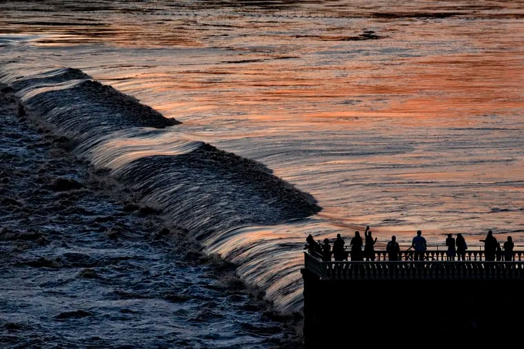 The sun sets on the above-flood-stage Schuykill as spectators on the Water Works watch the high, fast moving waters flow over the Schuylkill Dam Aug. 4, 2020 following a morning of heavy rain from Isaias.