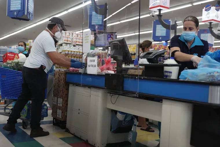 In this April 13 photo, Lay Guzman stands behind a partial protective plastic screen and wears a mask and gloves as she works as a cashier at the Presidente Supermarket in Miami