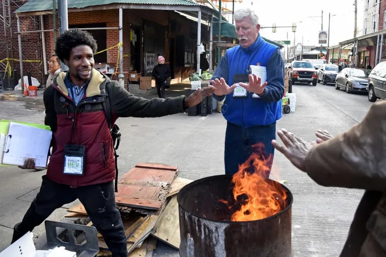Matthew Batte-Cooper (left) and Steve Horton stop to warm their hands at a barrel fire on South Ninth Street while doing outreach with Community Marketing Concepts. They were working as a beverage tax ground team, visiting corner stores and restaurants in the Italian Market.