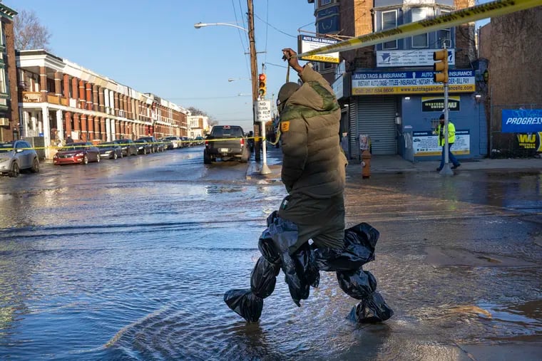 A neighborhood resident with makeshift waders made of garbage bags walks across the intersection of 56th Street at Chester, where water is flowing after a water main break. Philadelphia Water, Fire, and Police Departments were at a water main break on 56th Street between Springfield and Chester in Southwest Philadelphia on Wednesday morning, Feb. 9, 2022.