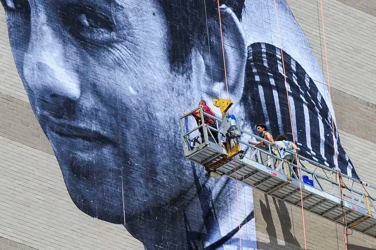 Artist JR, with fedora, installs his mural of Ibrahim, a Pakistani immigrant and Philadelphia resident, on the side of the Graham Building at 30 S. 15th St. The portrait is part of Mural Arts' "Open Source" series.
