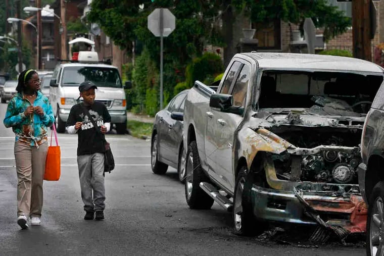 Pedestrians pass one of seven vehicles that were set ablaze early Wednesday on Georges Lane and West Berks Street in Wynnefield. A resident said an explosion woke her at 4:30 a.m.