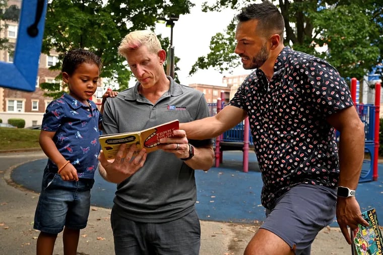 CJ Weiss (center) and Tyler Groll (right) read a book from the “Little Library “ with their 3-1/2 year-old son, Jackson Groll-Weiss, in their neighborhood park.