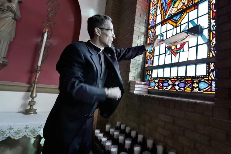 The Rev. Joseph Capella lifts cardboard that patched over a stained-glass panel broken by a vandal Jan. 6 at St. Lawrence Church in Lindenwold.
