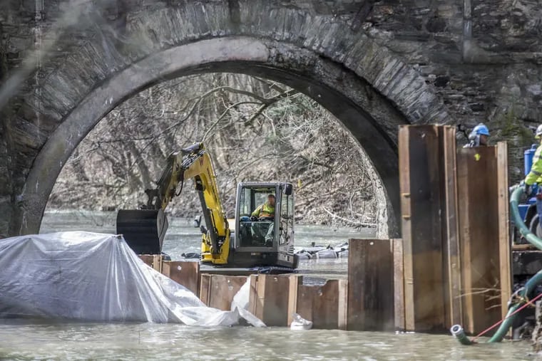 A backhoe works to add rocks and gravel to the creek bed, in order to level the bed, under the main arch of the Frankford Avenue Bridge, on March 13, 2018, as metal barriers are in place to keep more creek water from entering under the arch. That water is being pumped out so workers will be able to repair the bridge from underneath.