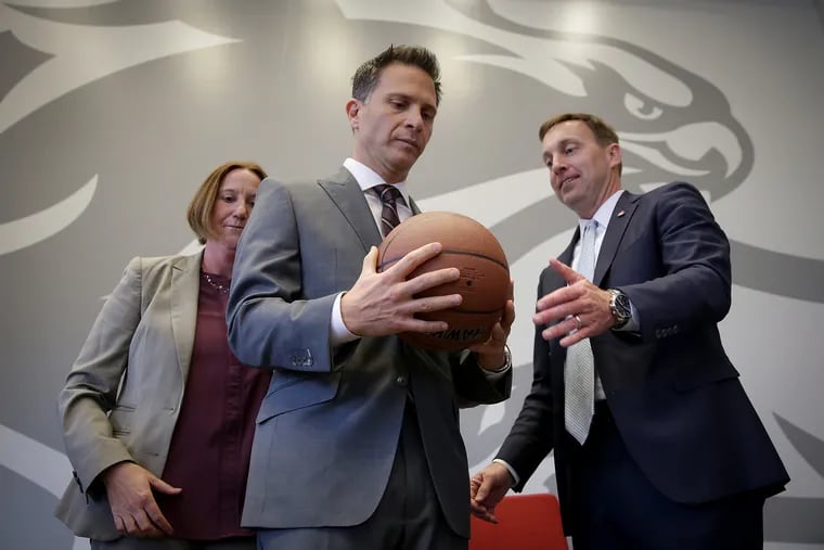 St. Joseph's University president Mark Reed (right) hands a basketball to new men's basketball coach Billy Lange at Lange's introductory press conference Thursday. Joining them on stage is athletics director Jill Bodensteiner.