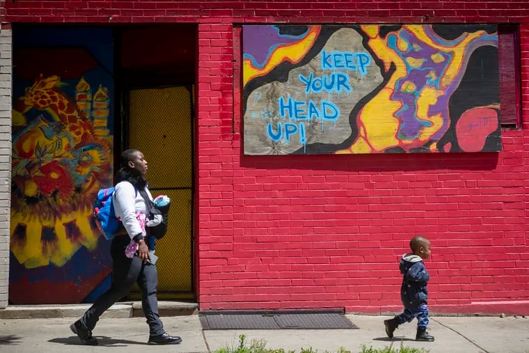 Jazmine Greene walks east along Lancaster Ave with her children on Thursday afternoon. Behind her is an abstract painting on plywood telling passersby to "Keep your head up!"
