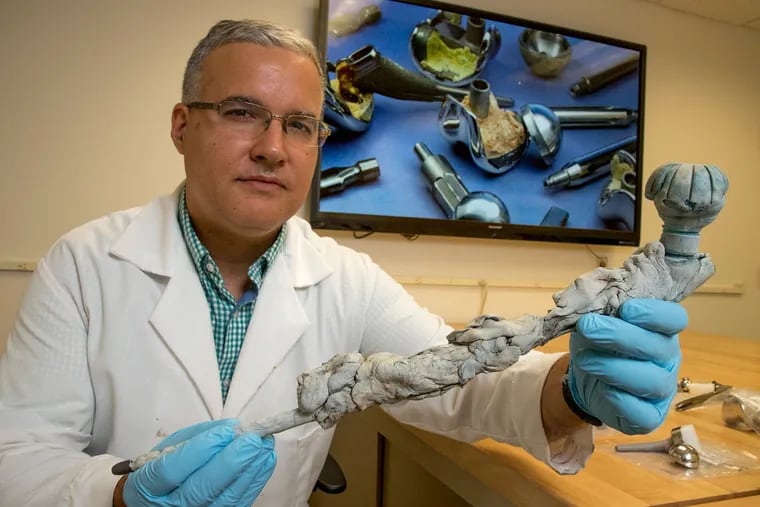 Steven Kurtz, director of the Implant Research Center at Drexel and a part time research professor, holds a hip anti-infection implant that was removed from a patient and sent to his center. On the screen behind him is a photo showing various failed implants.