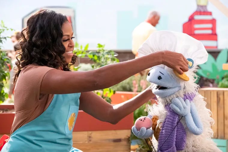 Michelle Obama puts a chef's hat on Waffles, a furry puppet with waffle ears and holding Mochi, a pink round puppet, on the set of the children's series "Waffles + Mochi."