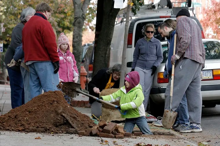 Joy Lawrence of the Pennsylvania Horticultural Society (center) cuts burlap from a maple as Ani Lesser, 9, helps with the digging at Aspen and 24th Streets in Philadelphia's Fairmount neighborhood. The region's TreeVitalize program is completing an effort to plant more than 14,000 trees this fall.