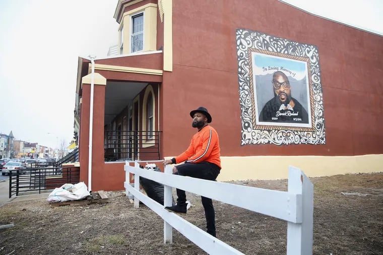Artist Ivben Taqiy stands for a portrait in front of his tribute mural to Derrick Rowland along North 52nd Street in West Philadelphia on Thursday, Feb. 15, 2018. Taqiy painted the mural in November for Rowland’s family.