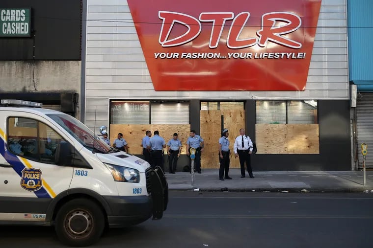 A small group of police stand outside the boarded-up DTLR store on 52nd Street near Market Street in West Philadelphia just after 6:45 p.m. on Monday, June 1, 2020. Many businesses along that stretch of 52nd Street were damaged on May 30 after property destruction and looting accompanied protests against the Minneapolis police custody death of George Floyd.