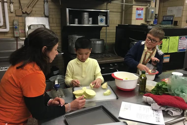 Teacher volunteer Lorrie Craley instructs Ivan Zheng and Travis Chopyak as they slice eggplant and grate cheese for eggplant parm at Comly Elementary.