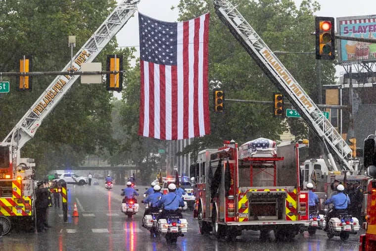 The body of Lt. Sean Williamson, Philadelphia Fire Department travels past American flag hung from Ladder trucks 14 and 19 outside Ladder 18 and Engine 59 on Hunting Park and 22nd Street. Fire Station Ladder 18, Engine 59 is where Lt. Williamson was stationed. Photo from funeral for fallen fire fighter,  Lt. Sean Williamson on Monday, June 27, 2022. His funeral mass is being held at Epiphany of Our Lord Church, 1121 Jackson St., Philadelphia.  Lt. Williamson died in the line of duty after a fire-damaged building collapsed in the Fairhill neighborhood, Philadelphia on June 18th.