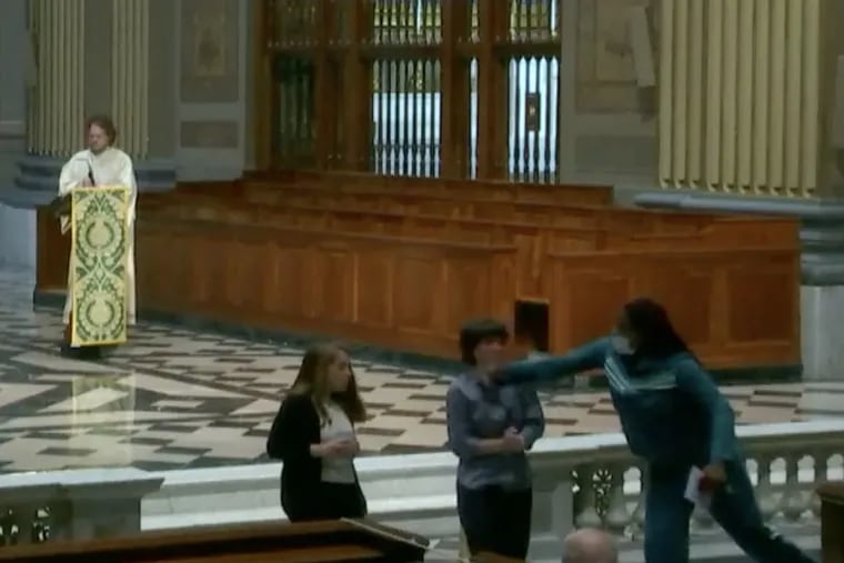 In this screenshot from the livestream of Sunday's Mass at the Cathedral Basilica of SS. Peter and Paul, a woman in a green track suit is shown punching a lector during the service. Philadelphia police said Monday that they had identified the suspect but had declined to pursue charges against her.