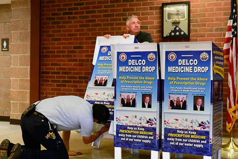 Vince Phillips (top), supplier, holds one 11 new “Delco Medicine Drop” boxes as Delaware County maintenence worker Paul O'Connell (on knees) readies the boxes for transportation to 10 Delaware County police departments and the Government Center in Media October 8, 2013. This followed press conference by the Delaware County Police Chiefs Association, Delaware County Council, District Attorney Jack Whelan, and the Delaware County Heroin Task Force unveiling the boxes which will be bolted to the floor at those sites, providing a place for citizens to drop off old medicines so they don't fall into the wrong hands. ( TOM GRALISH / Staff Photographer )