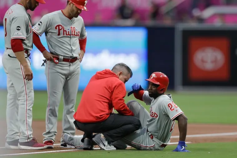 Philadelphia Phillies' Andrew McCutchen, right, is helped by a trainer after being injured while trying to get back to first base during the first inning of a baseball game against the San Diego Padres, Monday, June 3, 2019, in San Diego. Phillies manager Gabe Kapler, left, watches alongside first base coach Paco Figueroa. (AP Photo/Gregory Bull)