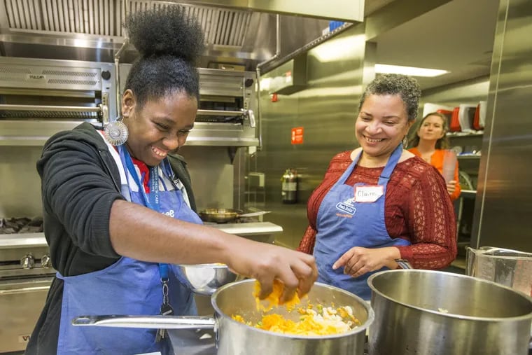 Christina Duckett-Brooker, left, adds grated cheddar to a pot of macaroni and cheese crafted by students in Cooking with Confidence, a program created in partnership with the Free Library of Philadelphia's Culinary Literacy Center and the Philadelphia Autism Project designed to teach adults with autism basic cooking skills.